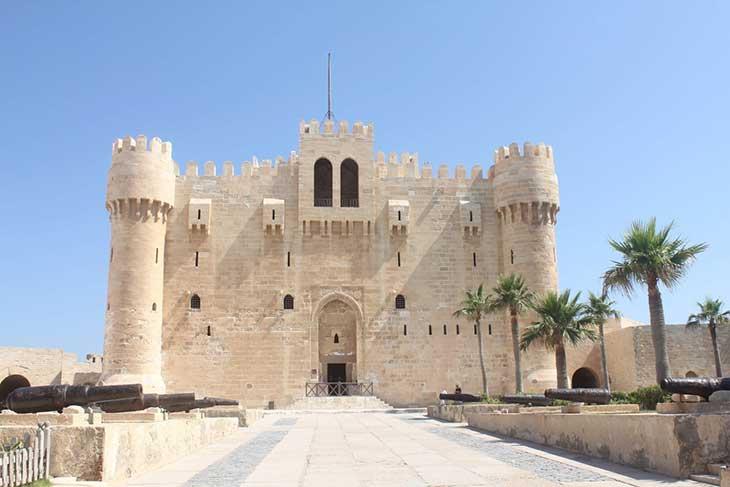 Front view of the historic Citadel of Qaitbay in Alexandria, Egypt, with palm trees and cannons surrounding the entrance.