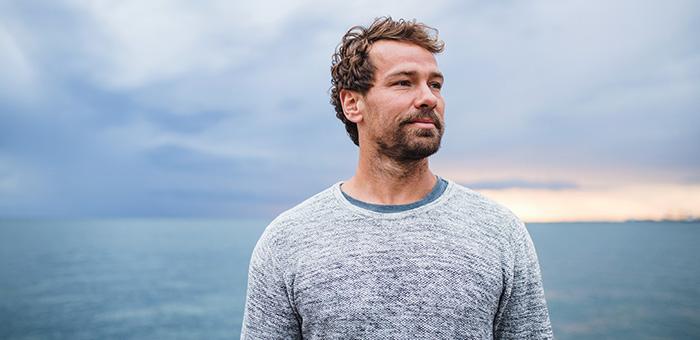 Man standing outdoors by the ocean, looking into the distance with a calm expression.