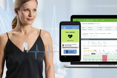 Woman wearing a wireless ECG monitoring device, with a smartphone and laptop displaying real-time heart data.